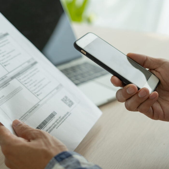 image of a person's hands, one with a bill and the other with a cell phone. there is a laptop in the distance.